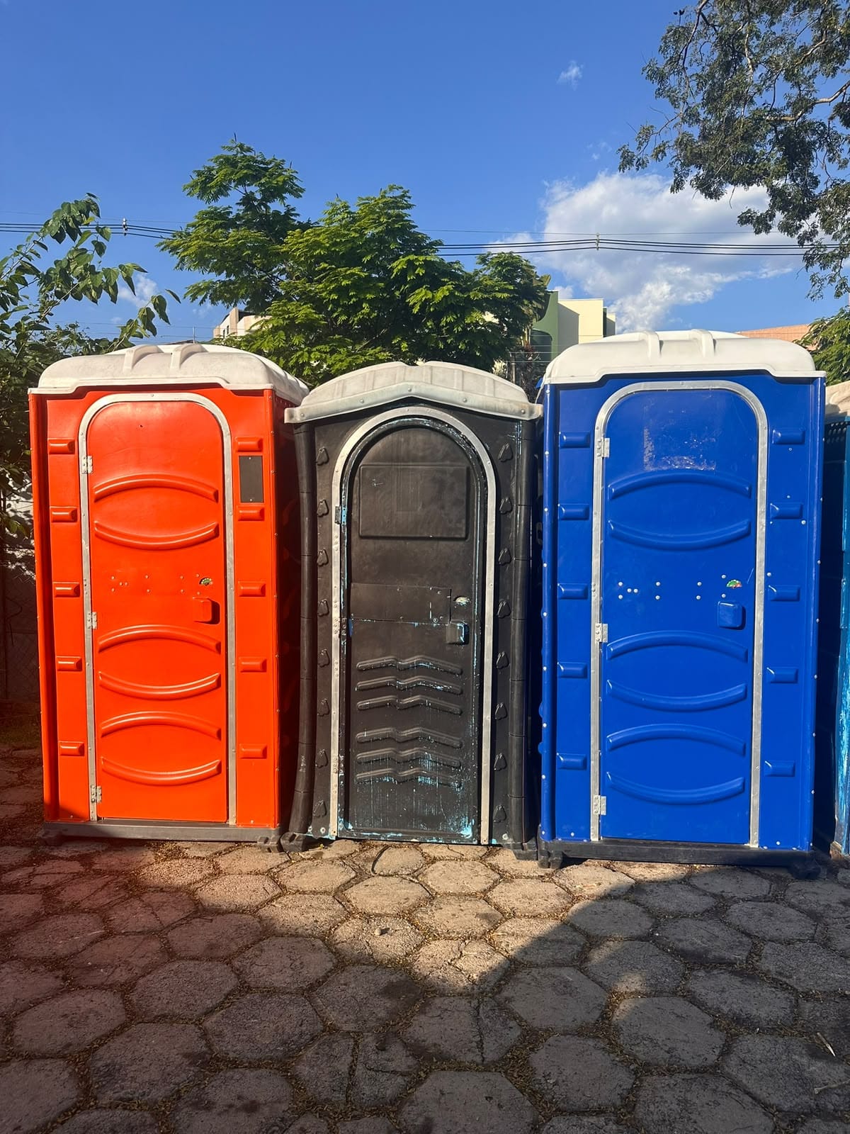 Três banheiros químicos, nas cores laranja, preta e azul, em piso de pedra